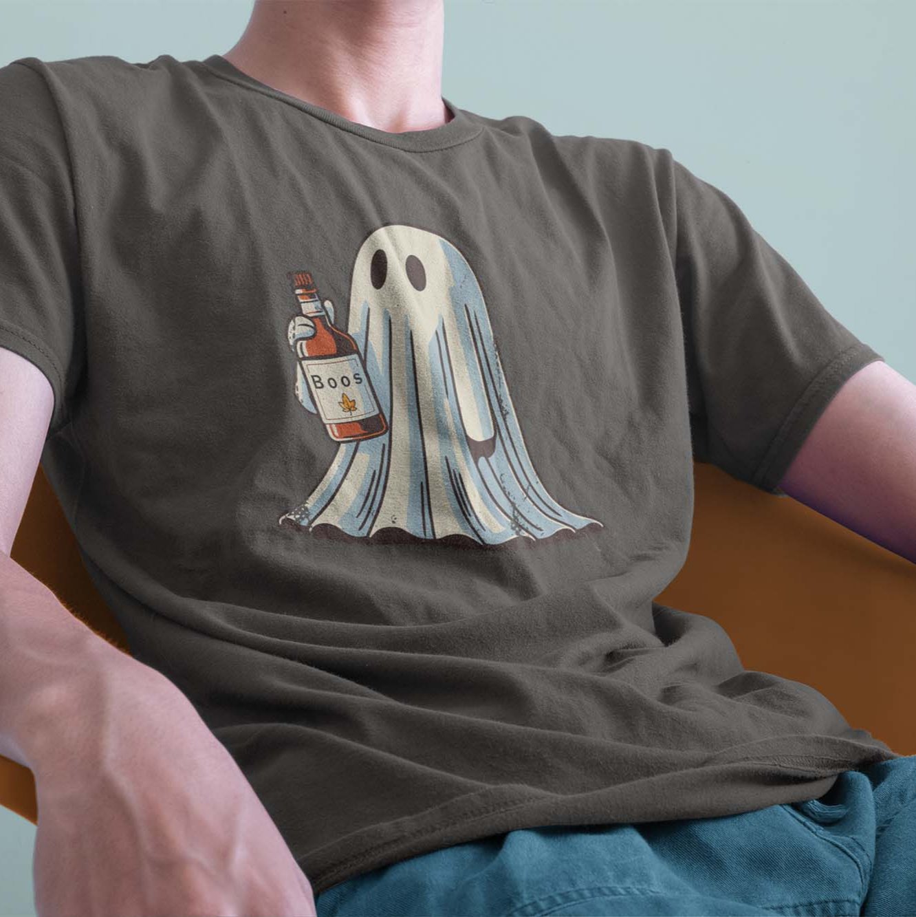 A male model leaning back on a chair, wearing a vintage Comfort Colors Tshirt in Pepper Gray color. The retro design features a cute bedsheet ghost holding a bottle of liquor that say Boos.