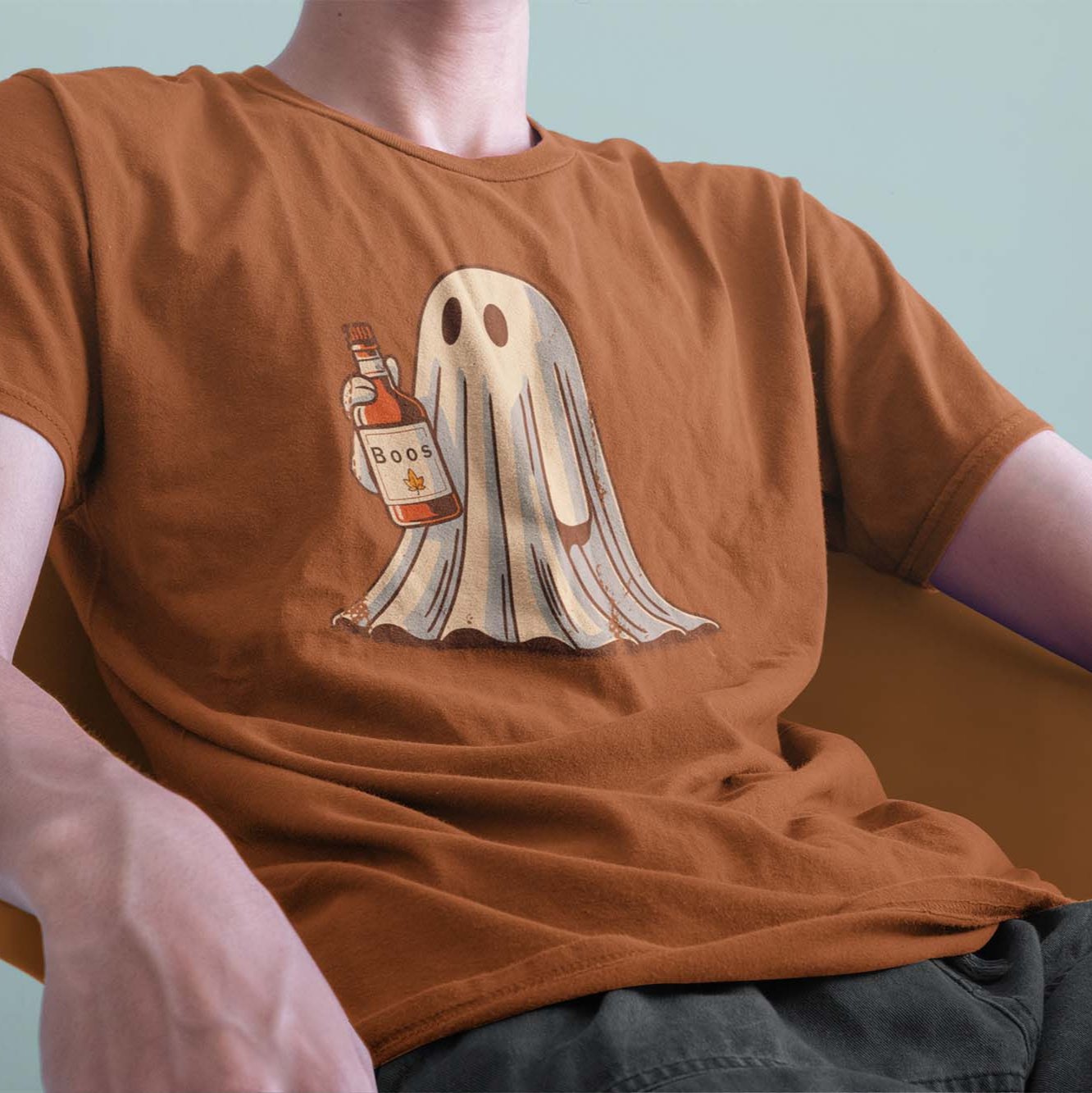 A male model leaning back on a chair, wearing a vintage Comfort Colors Tshirt in Yam Orange color. The retro design features a cute bedsheet ghost holding a bottle of liquor that say Boos.