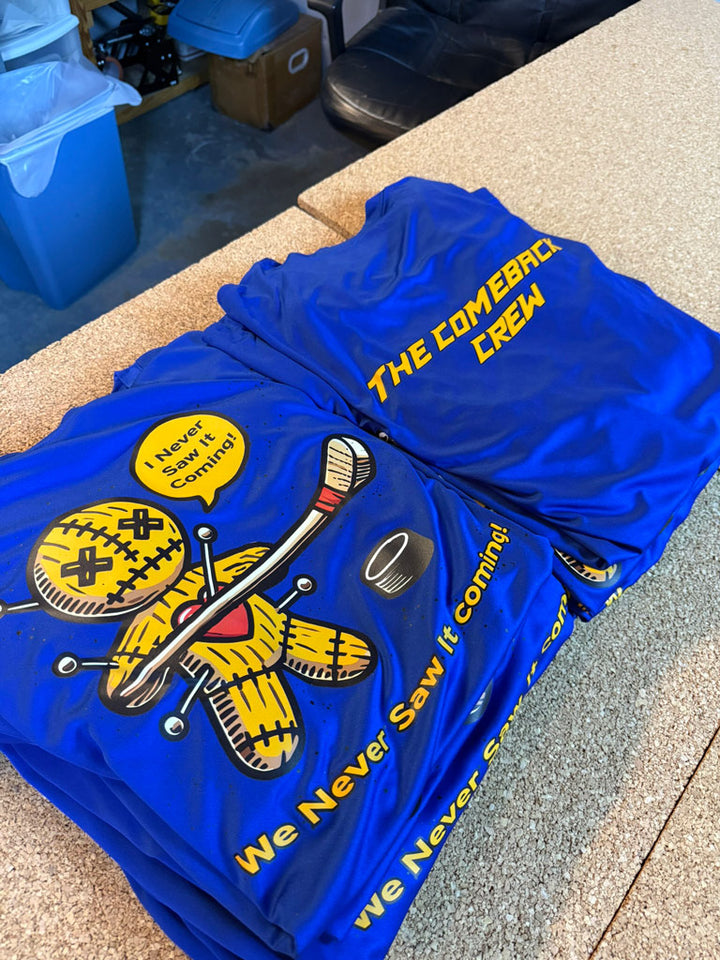 Stacks of royal blue custom crew jerseys printed front and back: back reads “The Comeback Crew,” front features a stitched voodoo puck graphic with “We Never Saw It Coming!”