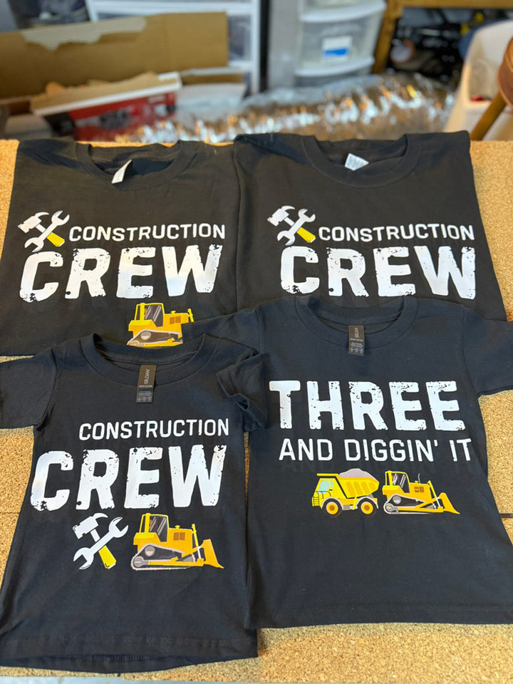 Black family set tees printed in white and yellow: two adult shirts read “Construction Crew” with tool and bulldozer icons; toddler shirt says “Construction Crew”; baby tee reads “Three and Diggin’ It” with dump truck graphic.