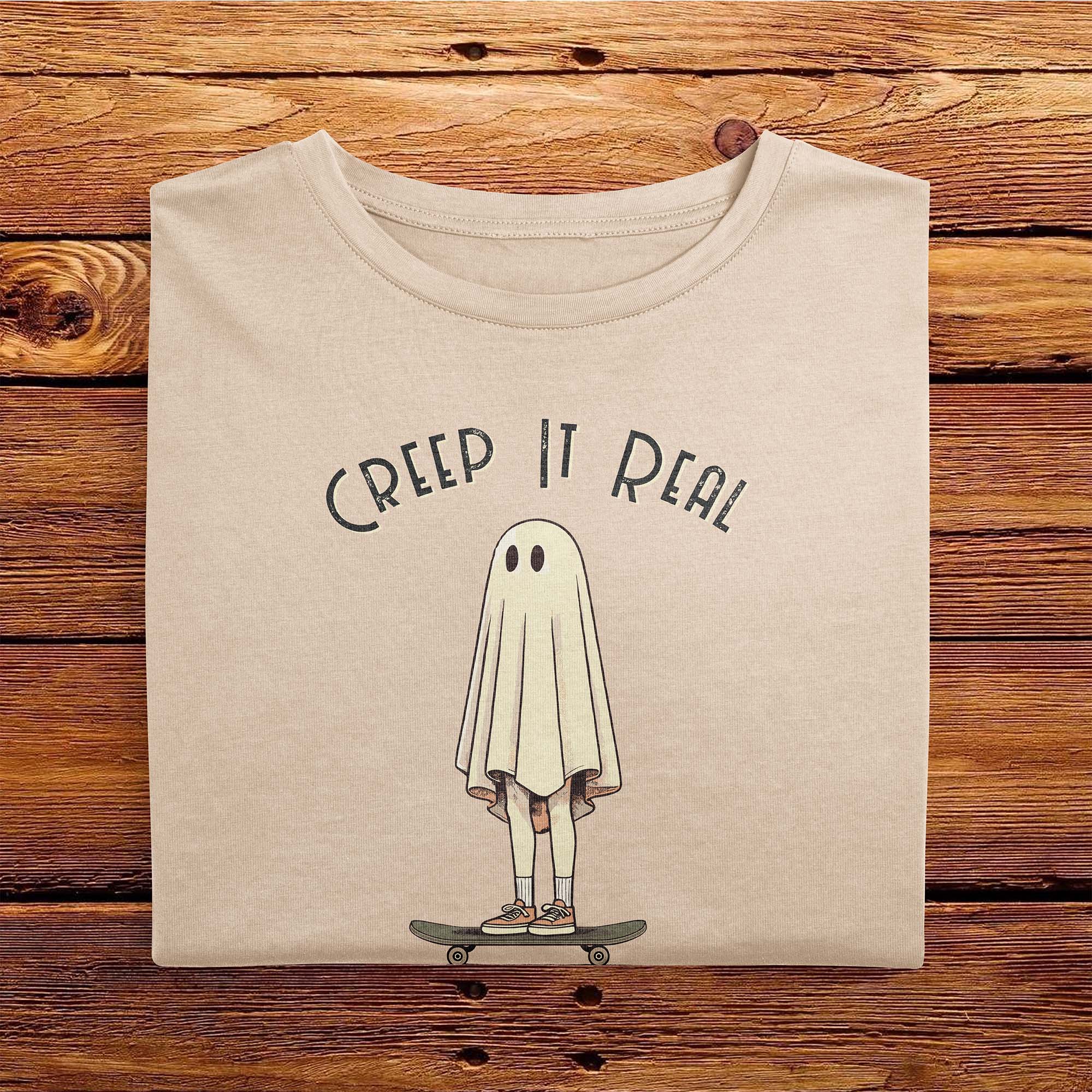 A folded Comfort Colors Tshirt in Ivory White color laying on a wooden table. The vintage design features a guy in a spooky bedsheet ghost costume standing on a skateboard, with text above him that says Creep It Real.
