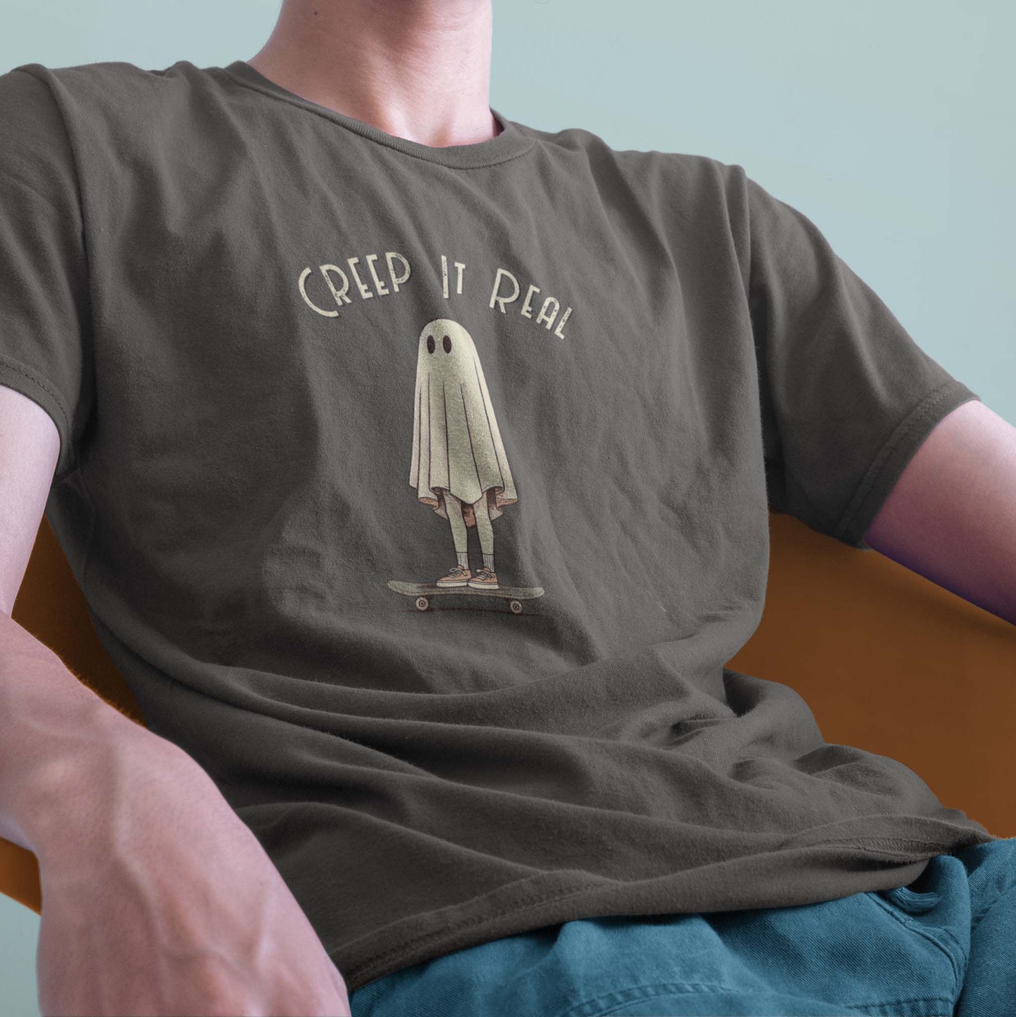 A male model leaning back in a chair wearing a Comfort Colors Tshirt in Pepper Gray. The vintage design features a guy in a spooky bedsheet ghost costume standing on a skateboard, with text above him that says Creep It Real.