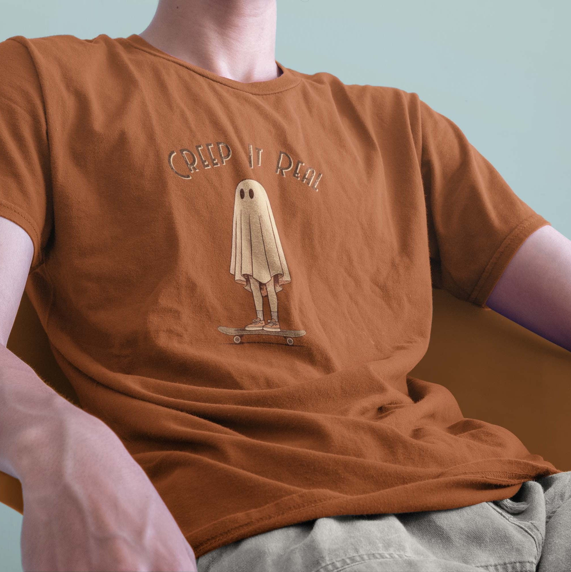 A male model leaning back in a chair wearing a Comfort Colors Tshirt in Yam Orange. The vintage design features a guy in a spooky bedsheet ghost costume standing on a skateboard, with text above him that says Creep It Real.