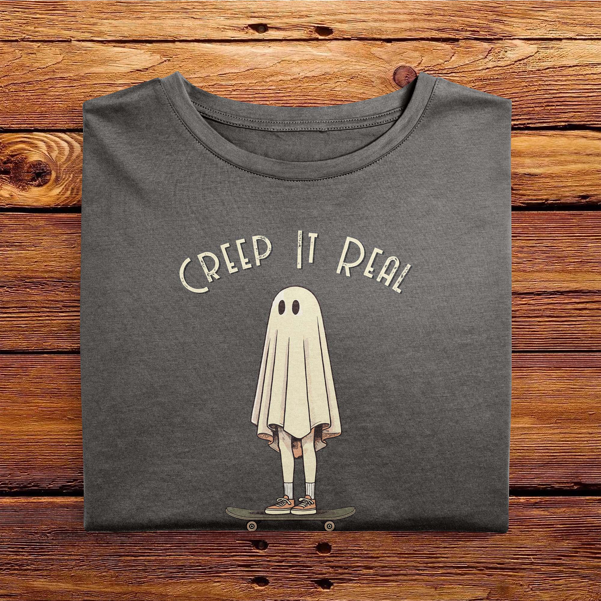 A folded Comfort Colors Tshirt in Pepper Gray color laying on a wooden table. The vintage design features a guy in a spooky bedsheet ghost costume standing on a skateboard, with text above him that says Creep It Real.