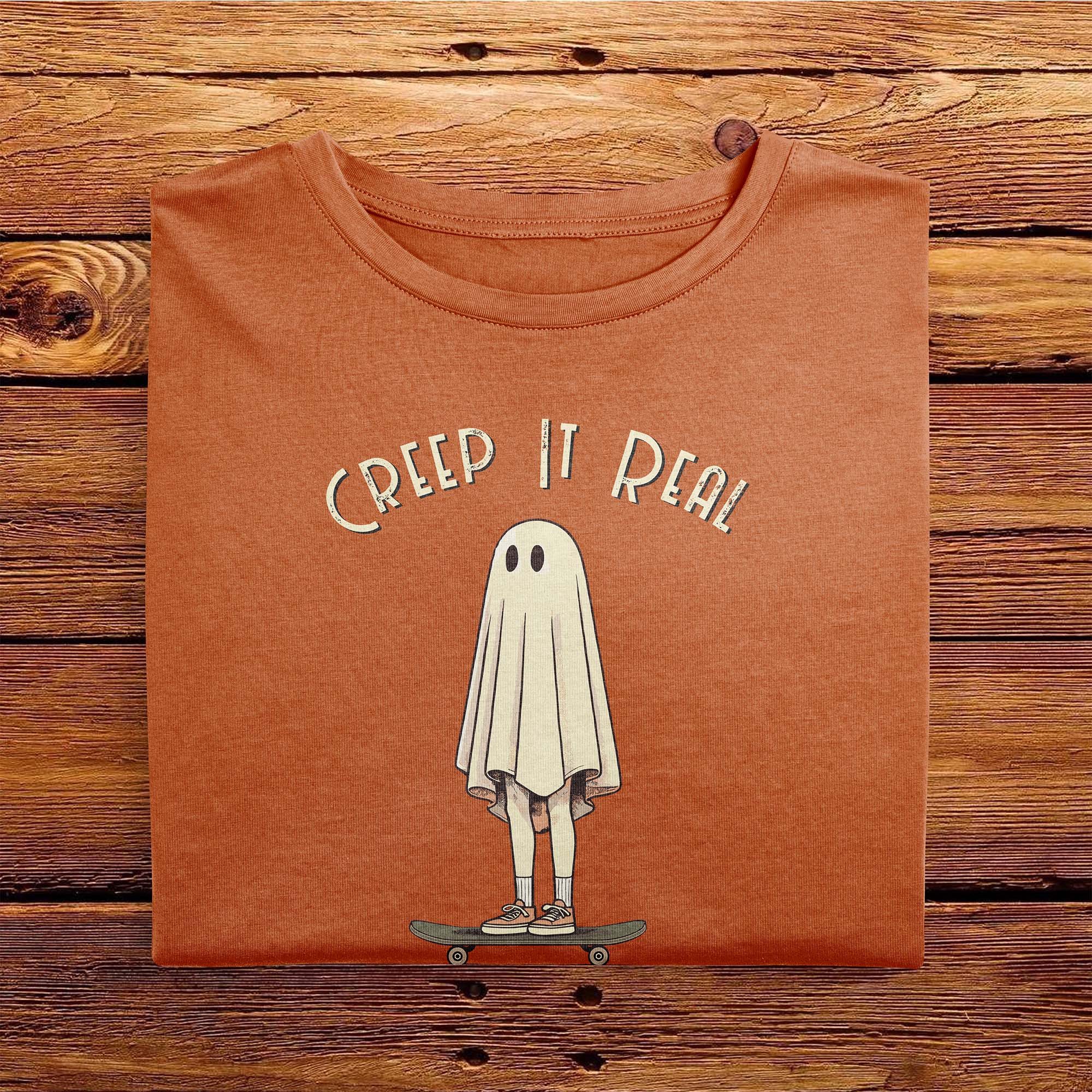 A folded Comfort Colors Tshirt in Yam Orange color laying on a wooden table. The vintage design features a guy in a spooky bedsheet ghost costume standing on a skateboard, with text above him that says Creep It Real.