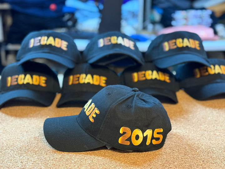 Black baseball caps stacked on a table, each embroidered “DECADE” in metallic gold, with one cap in front showing “2015” in gold on its side panel.