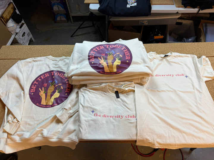 Cream-colored tees and sweatshirts stacked on a table, printed with pastel “the diversity club” text; sweatshirts feature a circular “Better Together” emblem with diverse hands.