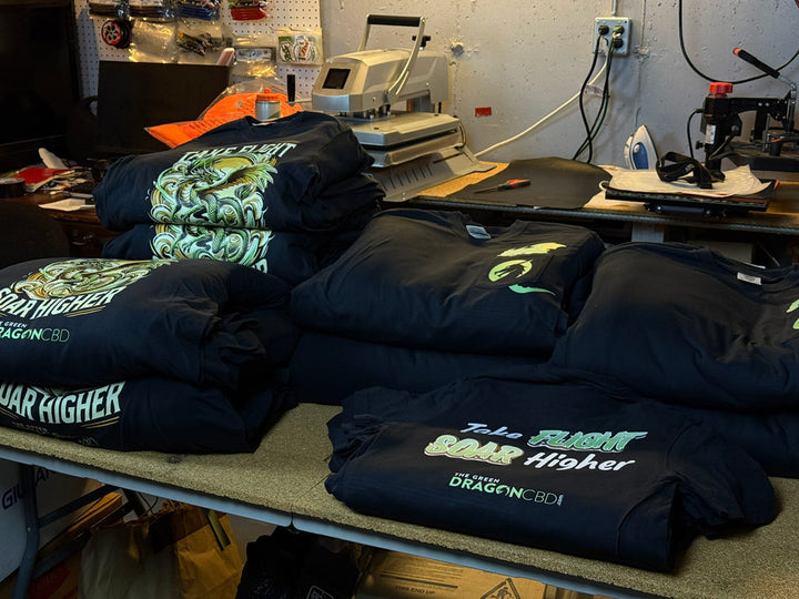 Stacks of black custom t-shirts on a workshop table featuring neon green dragon artwork and “Take Flight Soar Higher” text with The Green Dragon CBD branding under a heat-press machine.