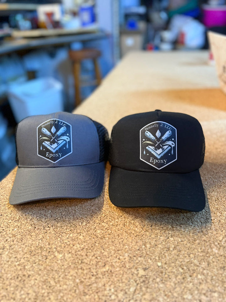 Two mesh-back trucker hats—one gray, one black—each printed with a white shield logo featuring trowels, epoxy drips, and “Donnelly J. LLC Epoxy” text.
