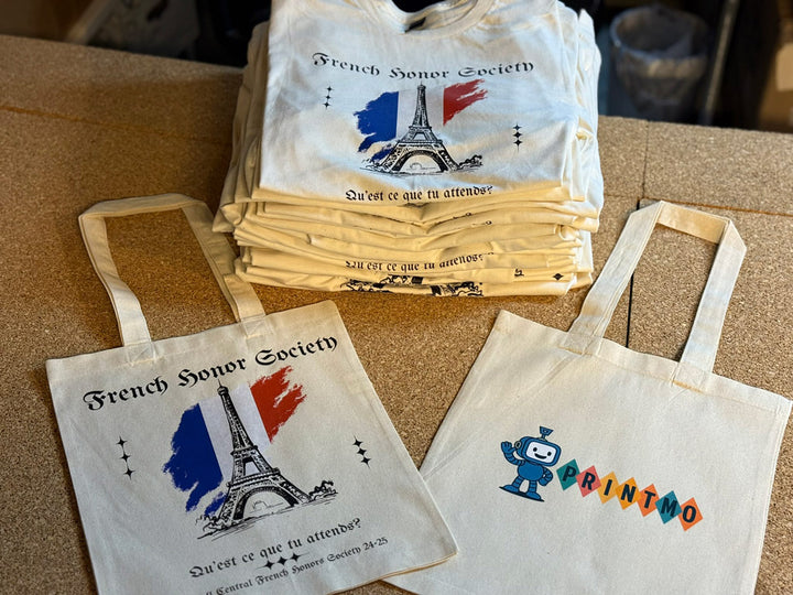 Stack of cream French Honor Society t-shirts printed with Eiffel Tower and tricolor brushstroke design, beside two matching canvas tote bags—one featuring the same France motif and one with the Print-MO robot logo.