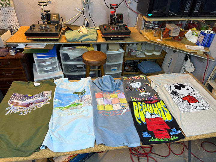 Six custom vintage-style tees laid flat on a workshop table: olive “Rock-Til-Ya-Tang 1971,” pale “Big Sur Coast,” pastel “Gettin’ The Funk Out,” plus three colorful Snoopy Peanuts designs.
