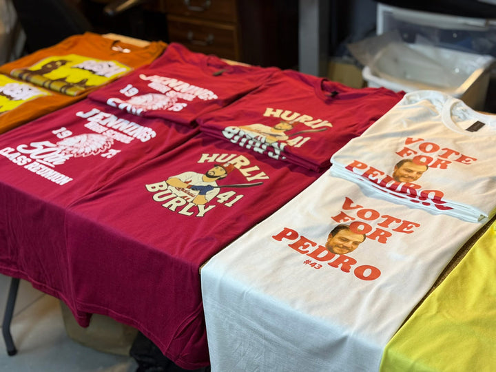 Flatlay of custom Print-MO tees: rust-orange DTF Rush concert shirt, maroon Jennings 50th reunion and Hurly Burly #41 designs, plus white Vote for Pedro shirts