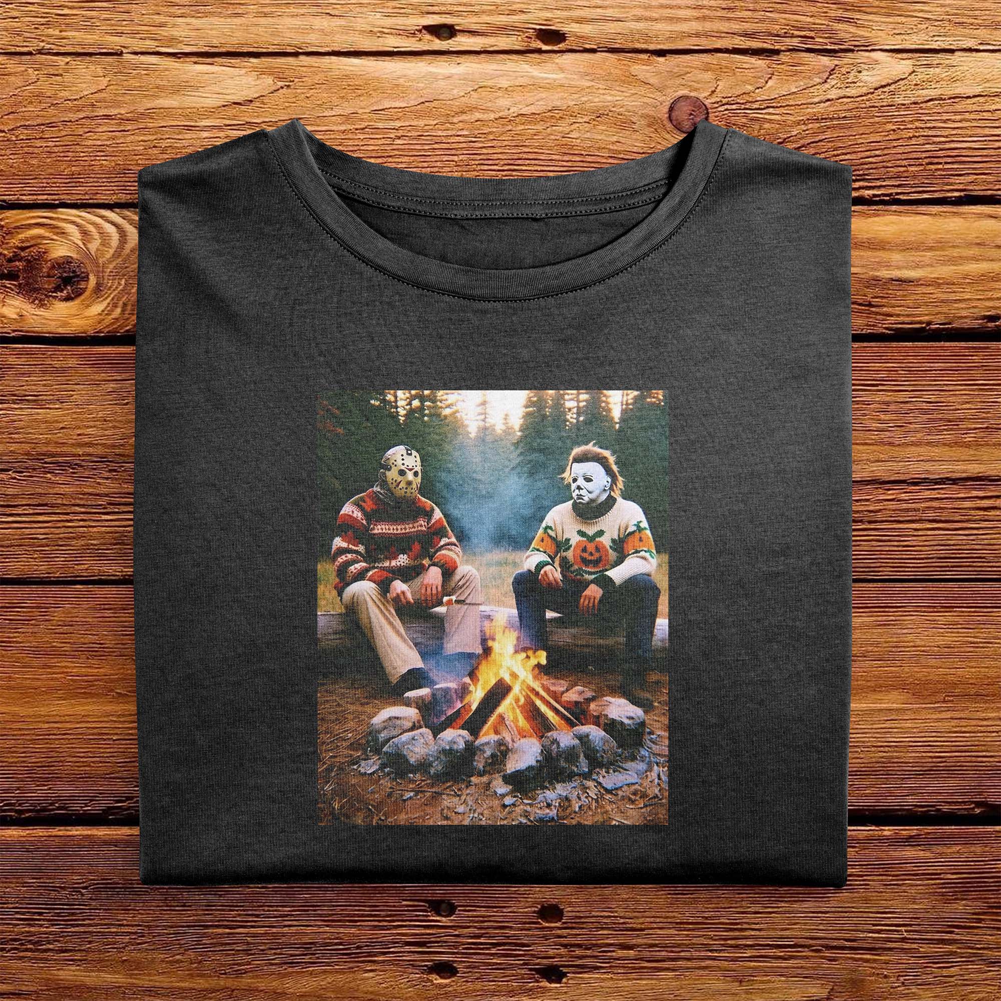 A Black folded Comfort Colors Tshirt, laying on a wooden table. The vintage design features Jason Vorhees from Friday the 13th, and Michael Meyers from Halloween, both in cozy sweaters sitting around a campfire roasting marshmallows.