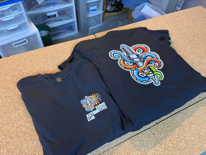 Two black KAIROS Automotive Club tees: chest print shows a spark plug wrapped in colorful octopus tentacles with starburst accents; back features a larger emblem.