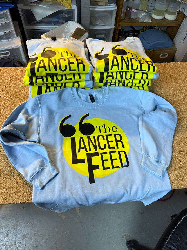 Light blue crewneck sweatshirts stacked and laid out, each printed with a bold yellow circle and black “The Lancer Feed” logo featuring stylized quotation marks above the text.