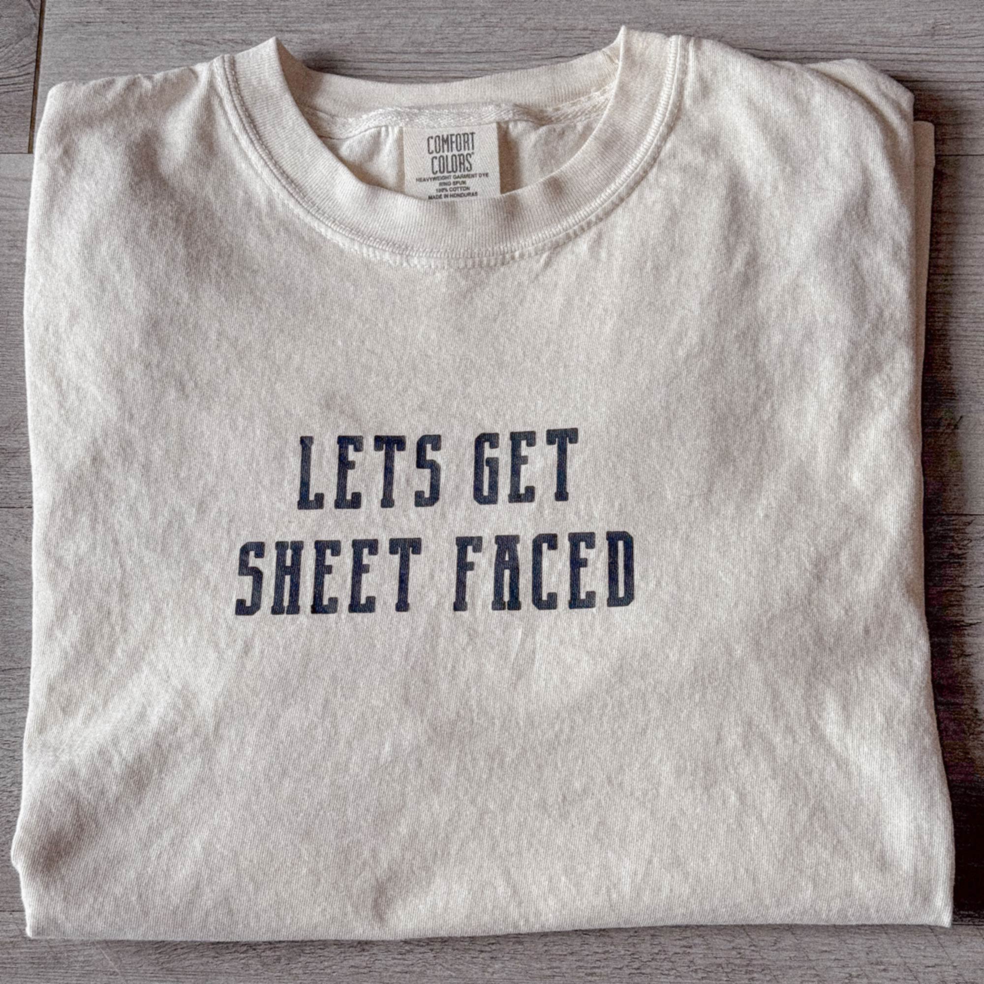 A Comfort Colors Tshirt in Ivory White color folded nicely with a modern wooden gray backdrop. There is a funny halloween themed pun on the front of the shirt that says Lets Get Sheet Faced.