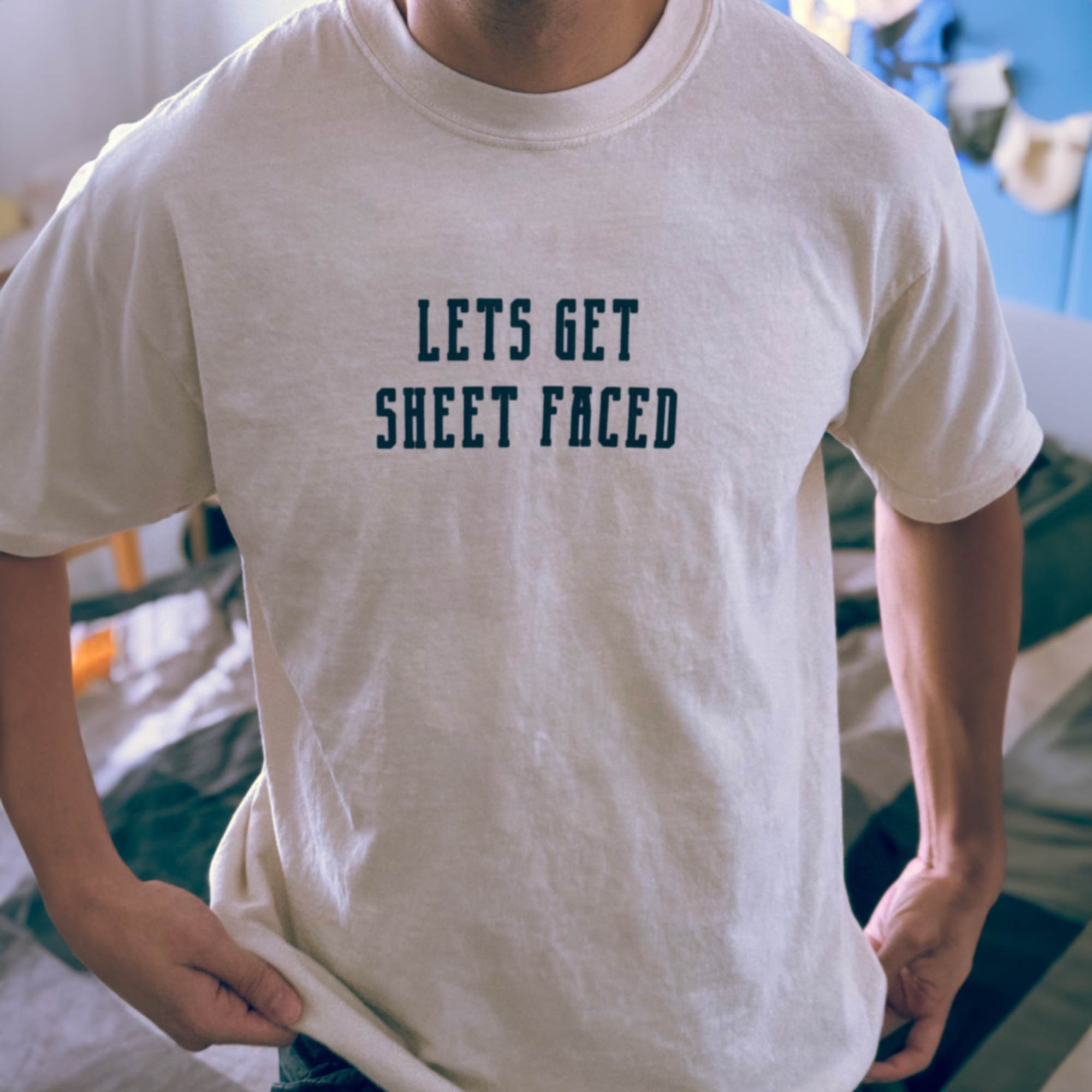 A male model wearing a Comfort Colors Tshirt in Ivory White color with his hands at his sides. There is a funny halloween themed pun on the front of the shirt that says Lets Get Sheet Faced.