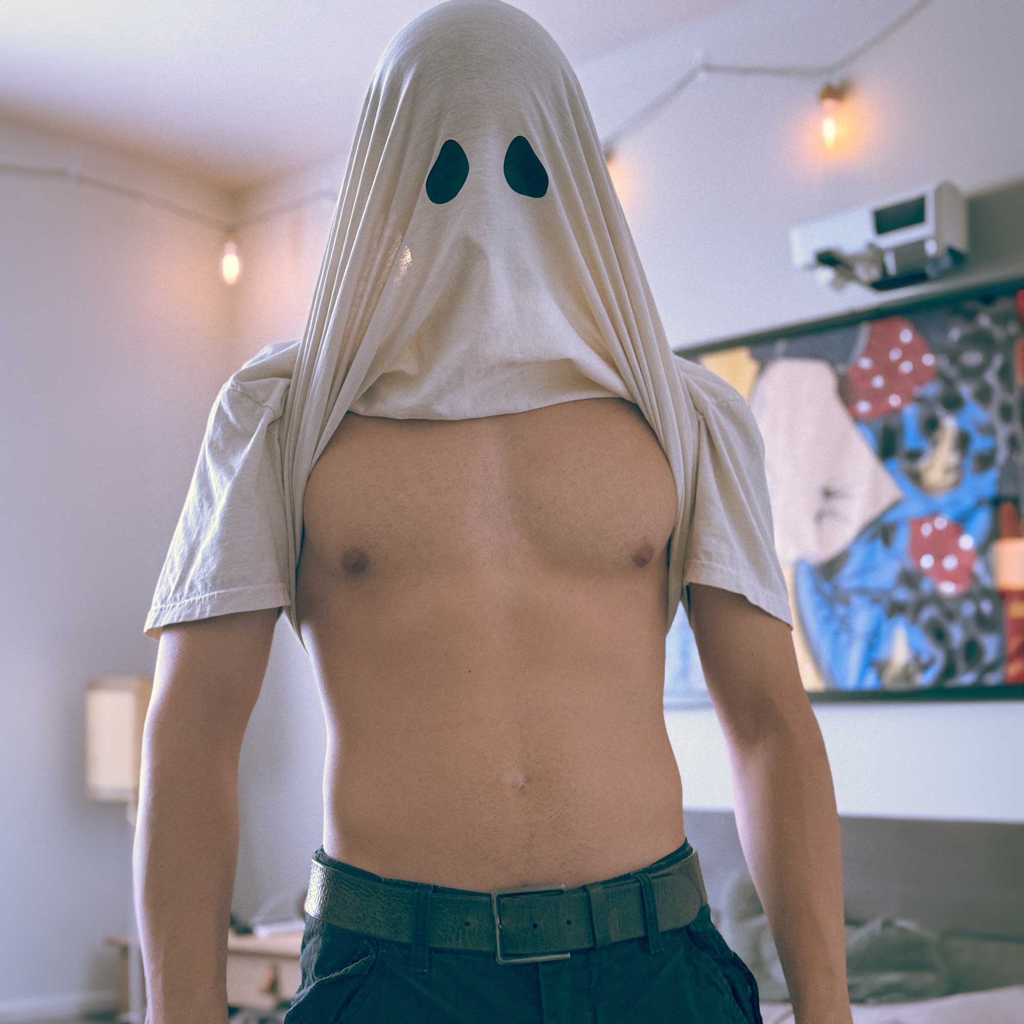 A male model wearing a Comfort Colors Tshirt in Ivory White. The shirt is pulled up above his head, revealing two black spots resembling the eyes of a bedsheet ghost. This shirt has a fun halloween themed design.