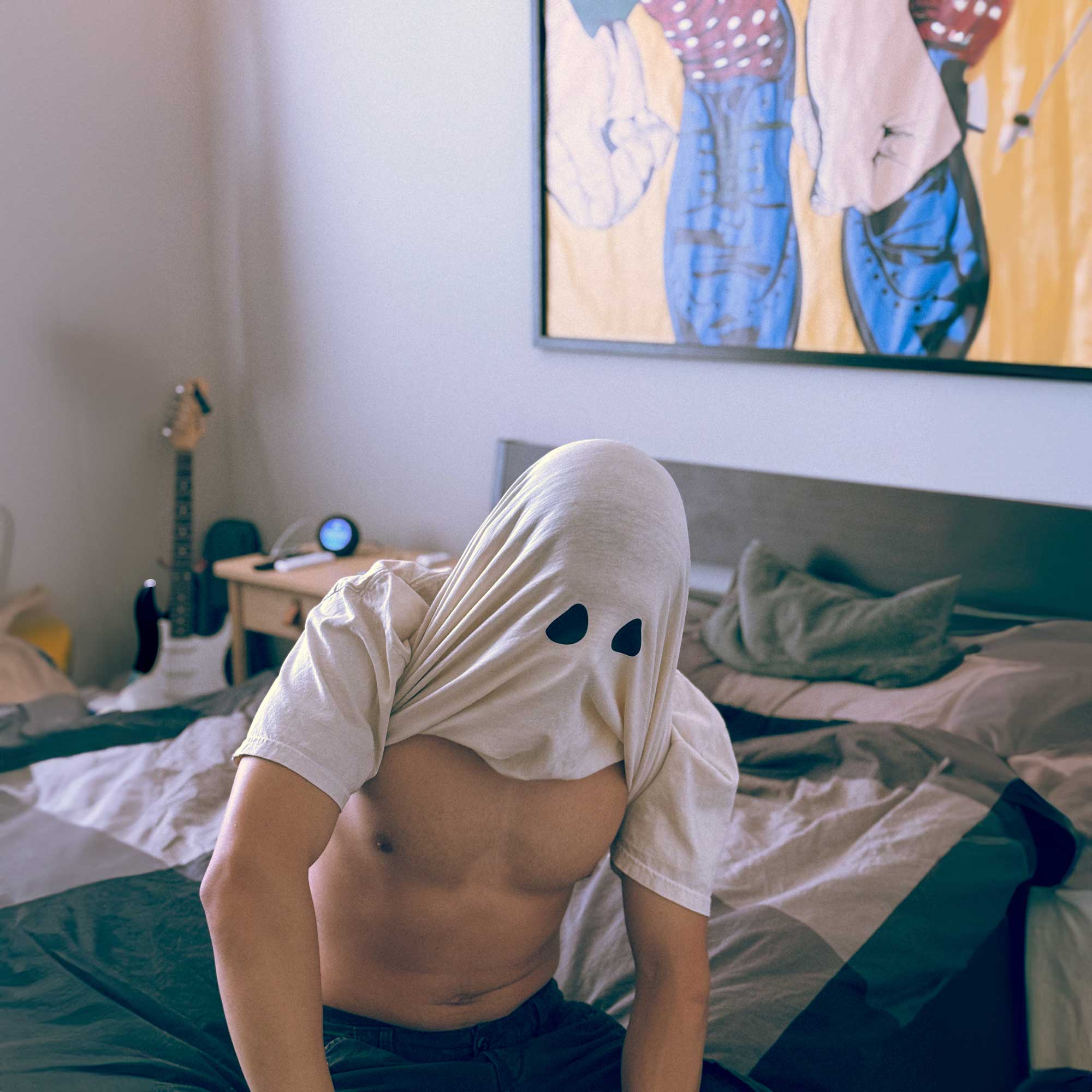 A male model wearing a Comfort Colors Tshirt in Ivory White sitting down on a bed. The shirt is pulled up above his head, revealing two black spots resembling the eyes of a bedsheet ghost. This shirt has a fun halloween themed design.