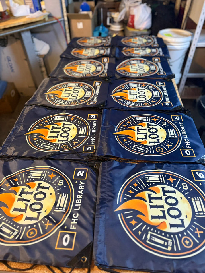 Navy drawstring backpacks numbered 1–8 laid out on a worktable, each featuring a circular “LIT LOOT” flame-and-book logo and FHC Library branding.