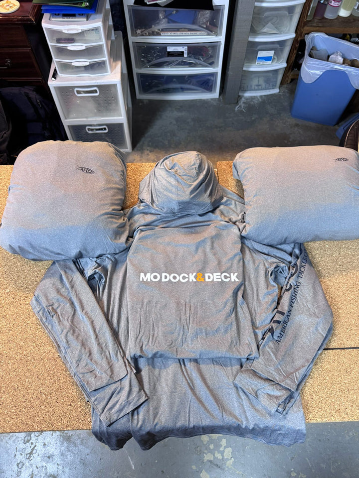 Gray performance hooded long-sleeve shirt laid flat between two matching pillow forms, printed with “MO DOCK & DECK” on the chest and “American Fishing Tackle Co.” down the sleeve.
