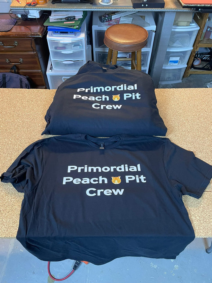 Black t-shirts stacked and laid flat on a workshop table, each screen-printed in white with “Primordial Peach Pit Crew” text across the chest for uniform crew apparel.