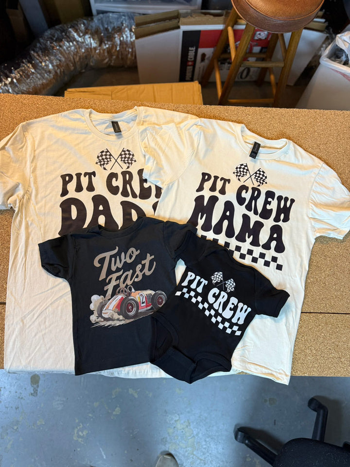 Cream-colored “Pit Crew Daddy” and “Pit Crew Mama” tees with checkered flag graphics, alongside a black “Two Fast” toddler tee and matching “Pit Crew” baby onesie.