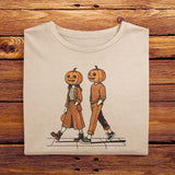 A folded vintage Comfort Colors Tshirt in Ivory White color, laying on a wooden table. The retro design features a cute couple walking together on a sidewalk, both wearing matching smiling jack-o-lanterns on their heads. Autumn Fall Vibes.