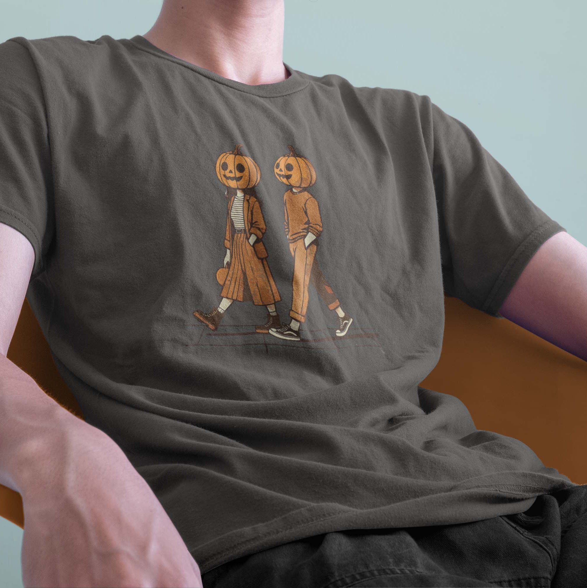 A male model leaning back in a chair wearing a Comfort Colors Tshirt in Pepper Gray. The retro design features a cute couple walking together on a sidewalk, both wearing matching smiling jack-o-lanterns on their heads. Autumn Fall Vibes.