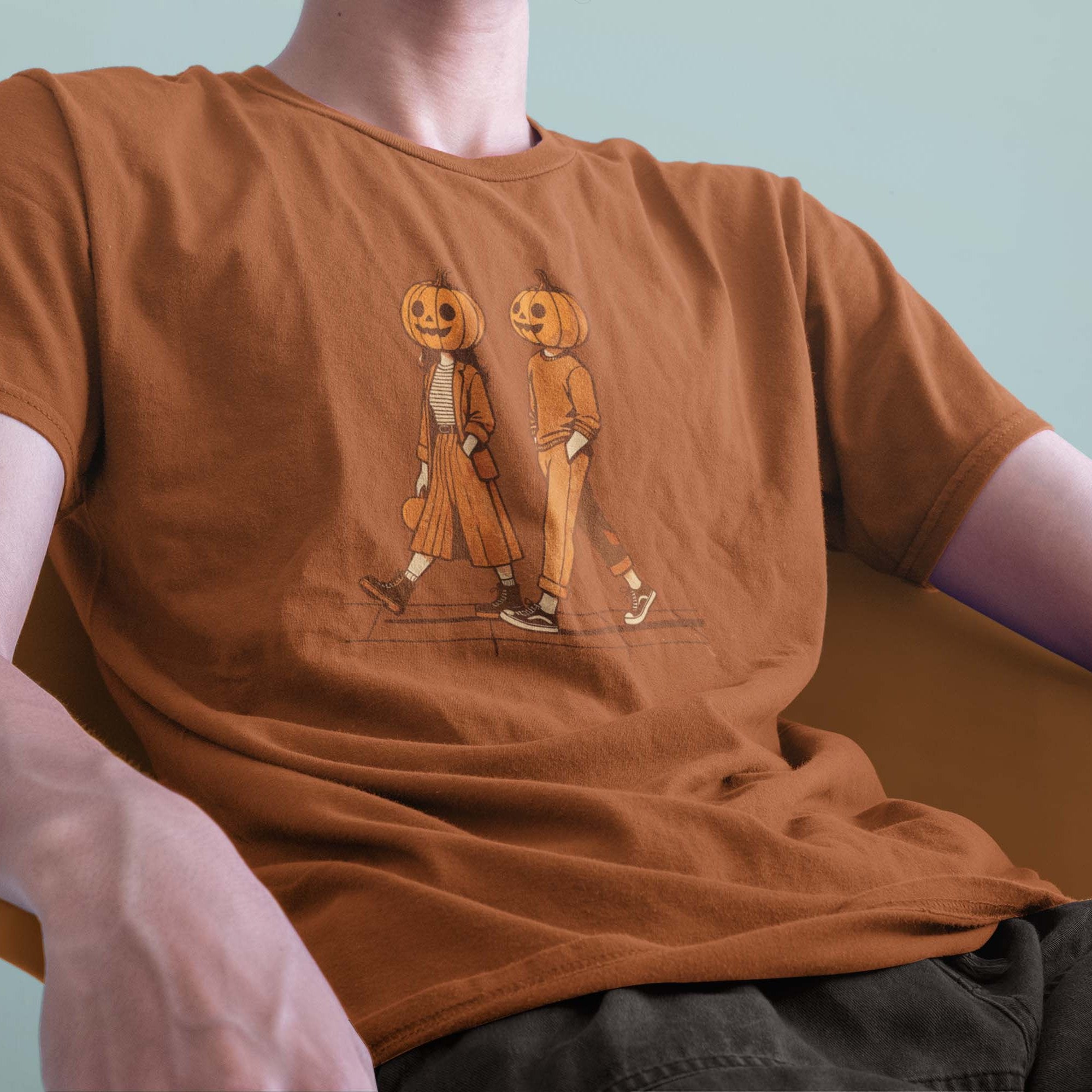 A male model leaning back in a chair wearing a Comfort Colors Tshirt in Yam Orange. The retro design features a cute couple walking together on a sidewalk, both wearing matching smiling jack-o-lanterns on their heads. Autumn Fall Vibes.