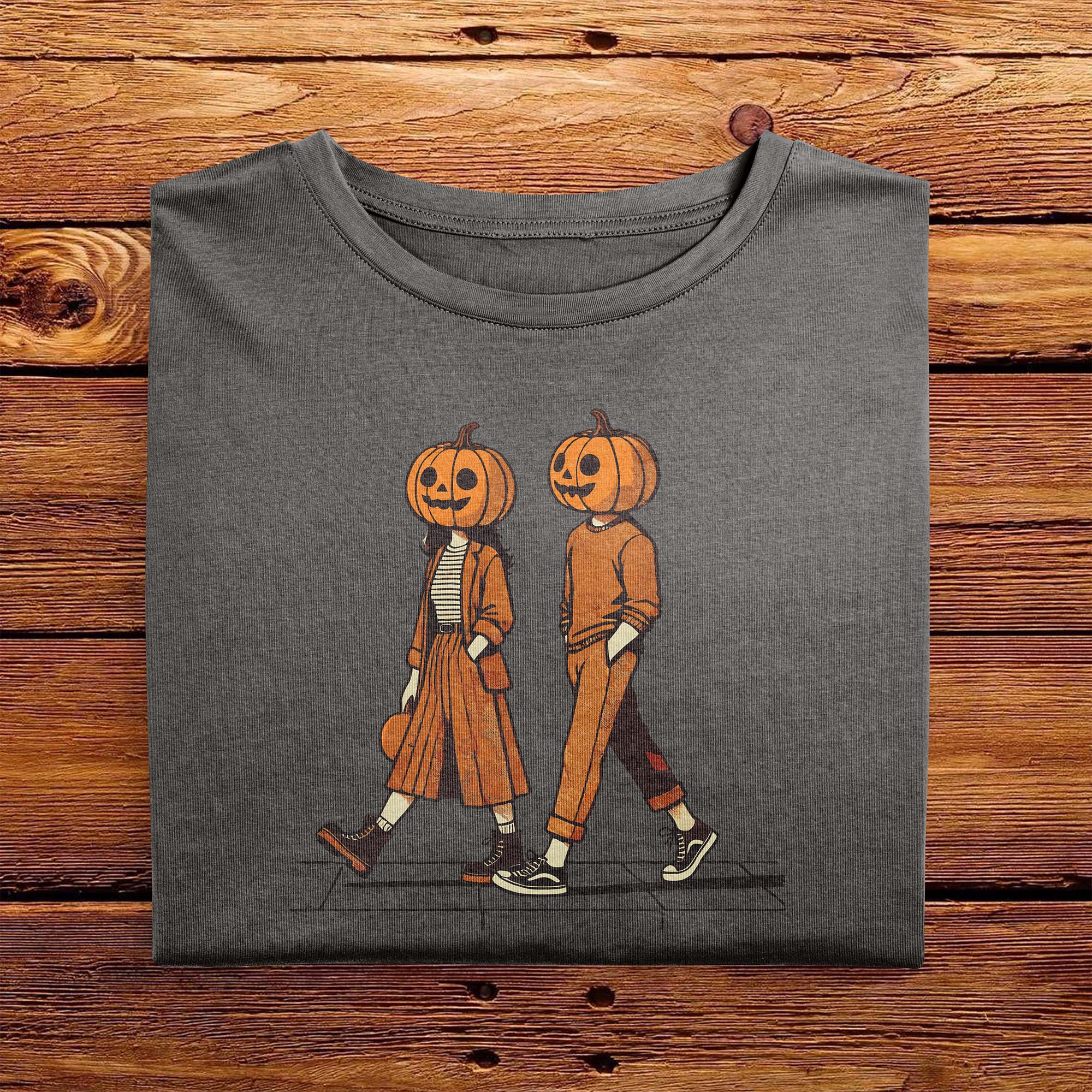 A folded vintage Comfort Colors Tshirt in Pepper Gray color, laying on a wooden table. The retro design features a cute couple walking together on a sidewalk, both wearing matching smiling jack-o-lanterns on their heads. Autumn Fall Vibes.