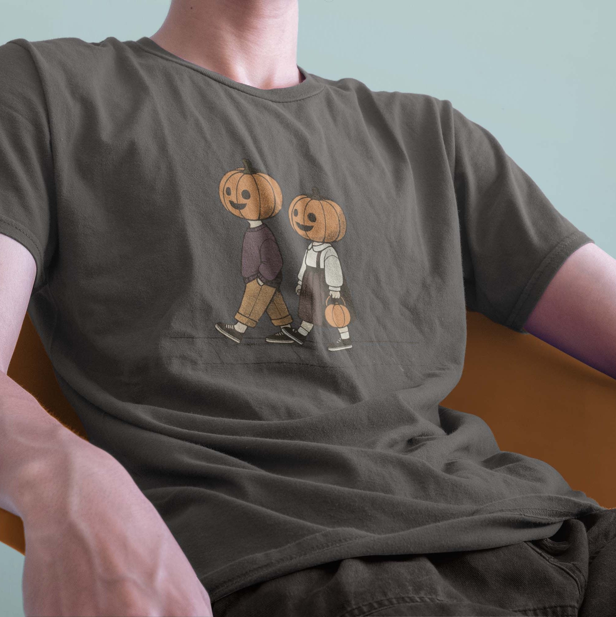 A male model leaning back in a chair wearing a Comfort Colors Tshirt in Pepper Gray color. The vintage design shows two young kids, a boy and a girl wearing fall apparel and pumpkins on their heads, strolling down a sidewalk