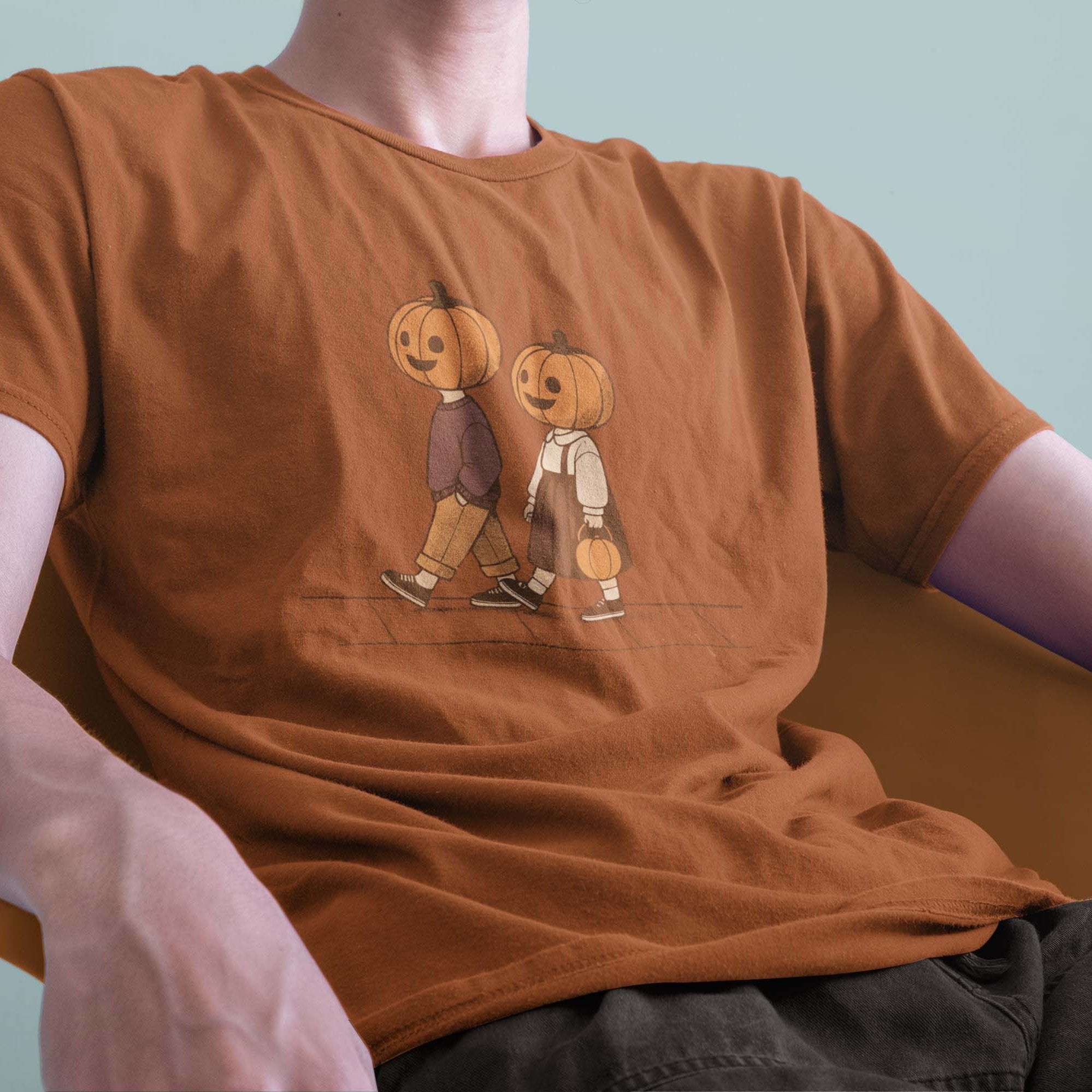 A male model leaning back in a chair wearing a Comfort Colors Tshirt in Yam Orange color. The vintage design shows two young kids, a boy and a girl wearing fall apparel and pumpkins on their heads, strolling down a sidewalk