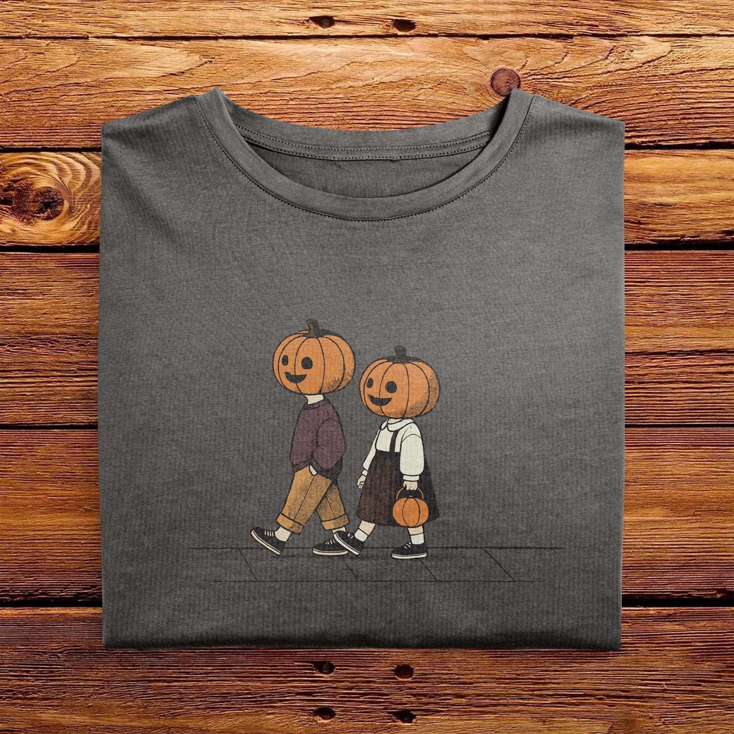 A folded Comfort Colors Tshirt in Pepper Gray color, laying on a wooden table. The vintage design shows two young kids, a boy and a girl wearing fall apparel and pumpkins on their heads, strolling down a sidewalk