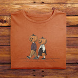 A folded Comfort Colors Tshirt in Yam Orange color, laying on a wooden table. The vintage design shows two young kids, a boy and a girl wearing fall apparel and pumpkins on their heads, strolling down a sidewalk