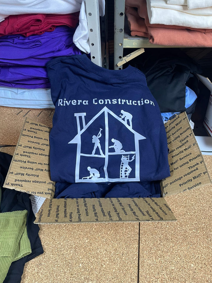 Navy Rivera Construction t-shirts with white house-shaped logo showing workers framing, roofing, plumbing, and tiling, branded apparel in a shipping box
