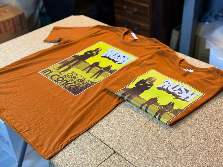 Two rust-colored custom t-shirts featuring a yellow-green vintage Rush concert poster print, one shirt laid flat and the other neatly folded on a workshop table.