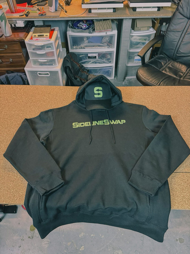 Black hoodie and matching cap displayed on a shop table, both printed with lime-green “SidelineSwap” text logo across the chest and front panel.