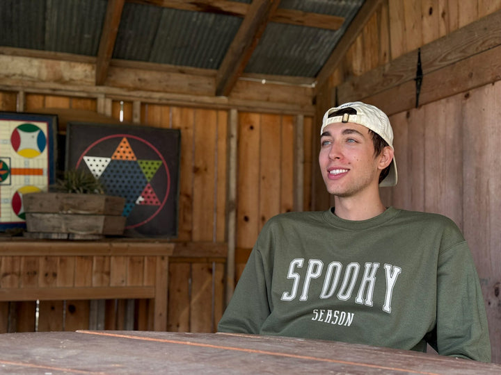 Olive green “Spooky Season” custom printed sweatshirt modeled in a rustic barn environment, highlighting high-quality fall apparel for seasonal fashion branding