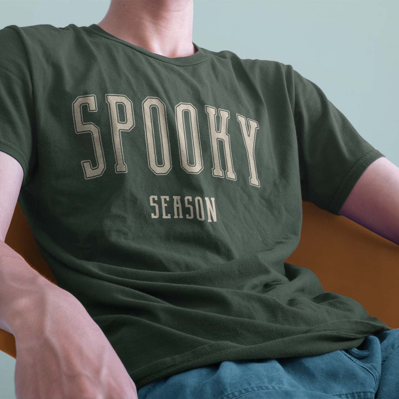 A closeup of a model in a chair wearing a comfort colors Tshirt in Moss Green color. The design includes off white text in a halloween font that says Spooky Season.