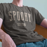 A closeup of a model in a chair wearing a comfort colors Tshirt in Pepper Gray color. The design includes off white text in a halloween font that says Spooky Season.