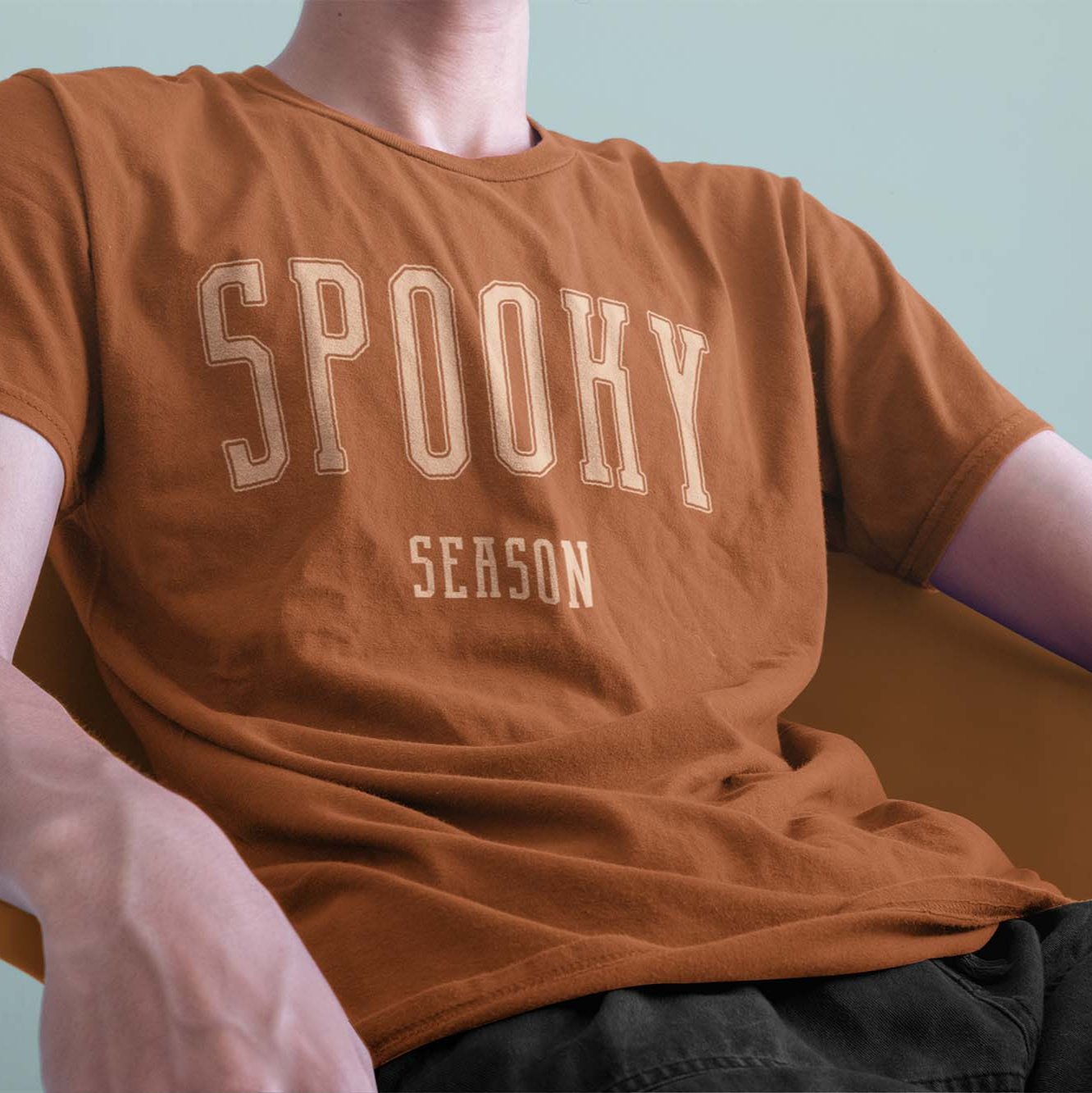 A closeup of a model in a chair wearing a comfort colors Tshirt in Yam Orange color. The design includes off white text in a halloween font that says Spooky Season.