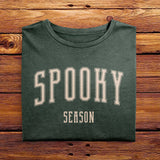 A folded Comfort Colors Tshirt in Moss green color, laying on a wooden table. The design includes off white text in a halloween font that says Spooky Season.