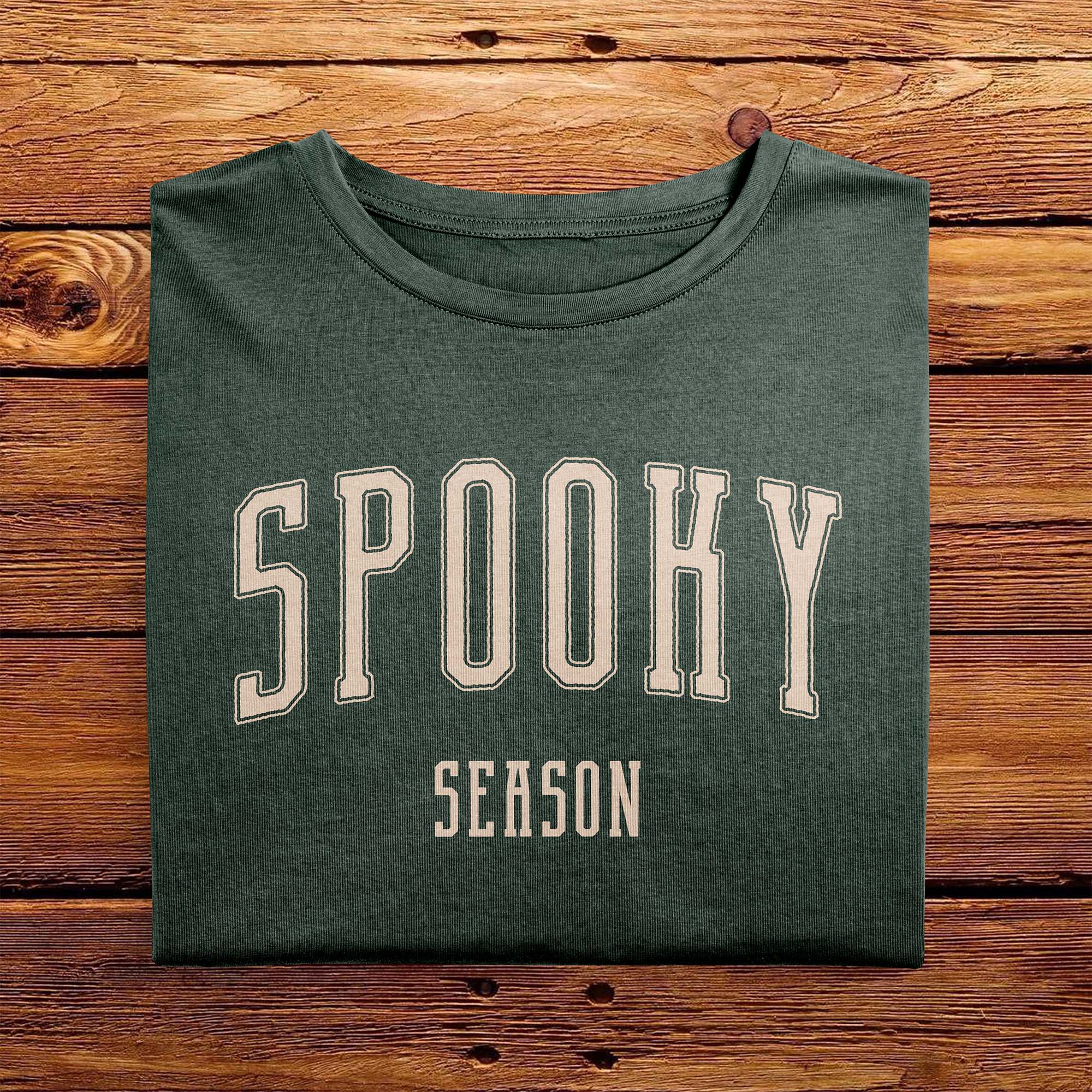 A folded Comfort Colors Tshirt in Moss green color, laying on a wooden table. The design includes off white text in a halloween font that says Spooky Season.