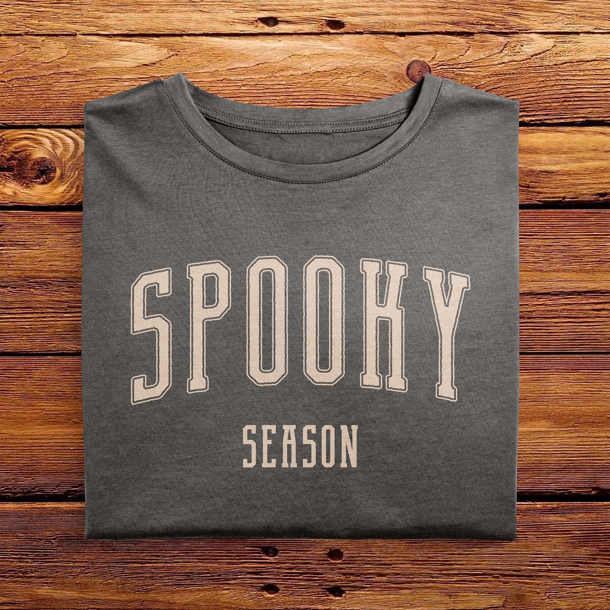 A folded Comfort Colors Tshirt in Pepper Gray color, laying on a wooden table. The design includes off white text in a halloween font that says Spooky Season.