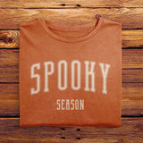 A folded Comfort Colors Tshirt in Yam Orange color, laying on a wooden table. The design includes off white text in a halloween font that says Spooky Season.