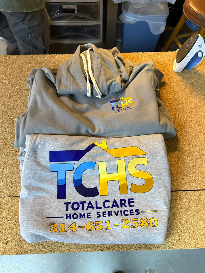 Stack of gray hooded sweatshirts printed with large TCHS Total Care Home Services logo and phone number 314-651-2580 across the front chest for branded apparel