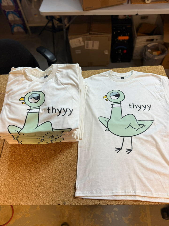 White t-shirts stacked and laid flat featuring a stylized green pigeon cartoon with puffed chest and the playful text “thyyy” alongside.