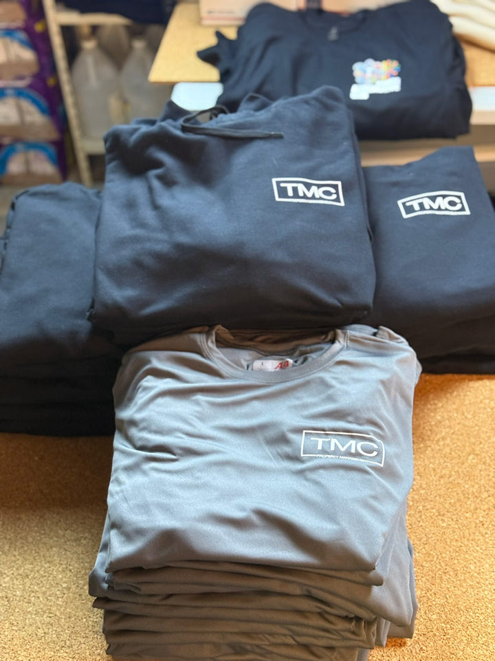 Stacks of black hoodies and tees and gray performance shirts on a table, each printed with a crisp white “TMC” logo inside a rectangular border.