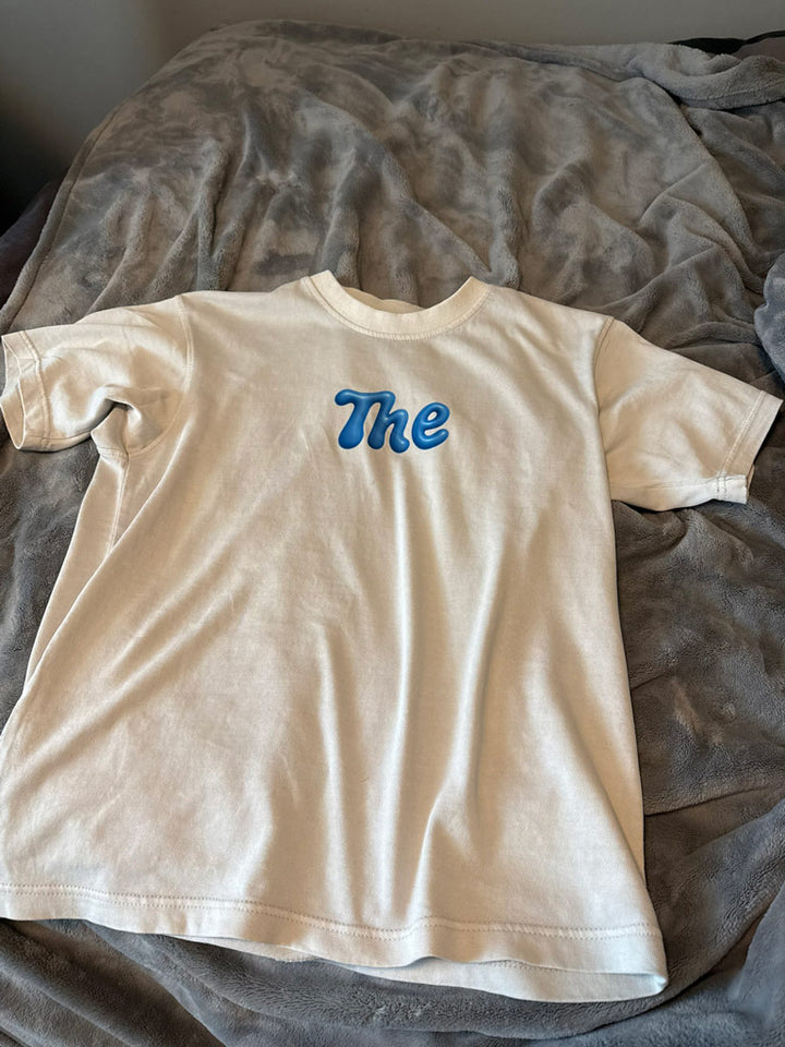 Beige t-shirt laid flat, printed with a shiny blue bubble-letter “The” centered on the chest for a playful minimalist design.